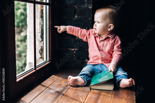 a little boy sits on the windowsill and holds a green book in his hands