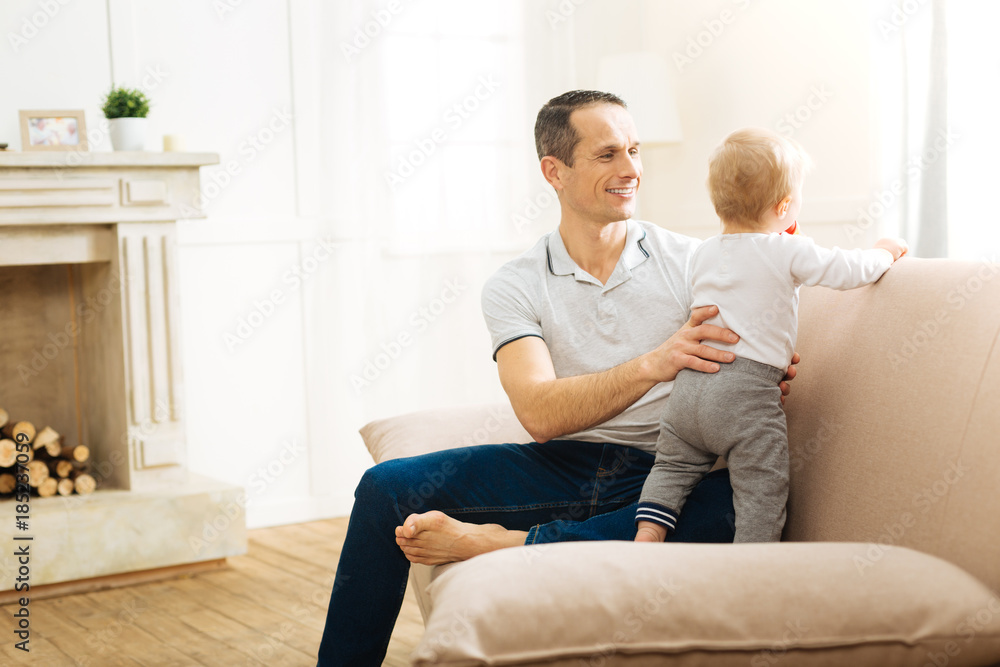 Pleasant timespending. Positive happy responsible father protecting his cute little baby from falling from the big sofa while touching his back and tummy and looking attentively at him