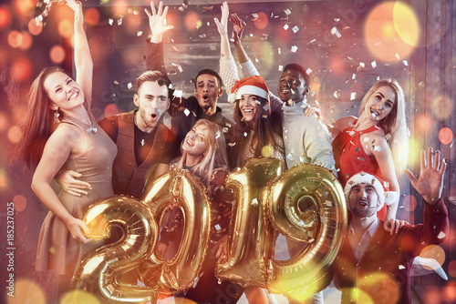 New 2019 Year is coming! Group of cheerful young multiethnic people in Santa hats carrying gold colored numbers and throwing confetti on the party