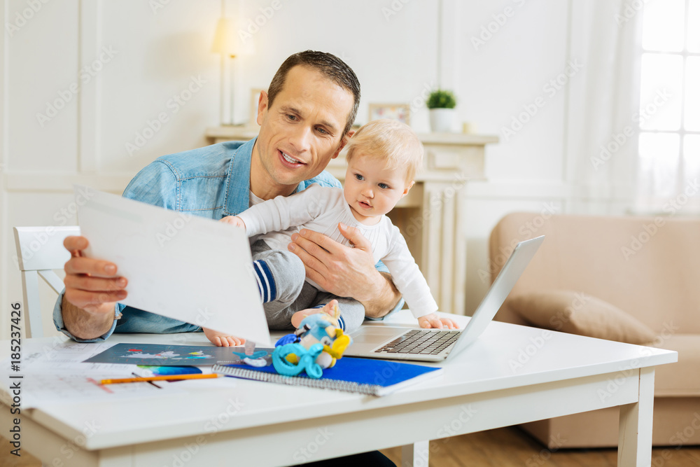 Strange information. Attentive clever young man feeling concentrated while sitting with his cute little baby and reading the notes on his project aloud Strange information. Attentive clever young man feeling concentrated while sitting with his cute little baby and reading the notes on his project aloud