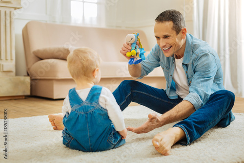 Bright toy. Attentive loving father feeling excited while sitting on the floor with a bright toy in his hand and attracting his childs attention while smiling and showing the new toy