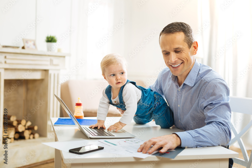 Exploring technologies. Cute pretty baby crawling on the table and touching a modern laptop while a kind calm father noticing it and smiling Exploring technologies. Cute pretty baby crawling on the table and touching a modern laptop while a kind calm father noticing it and smiling