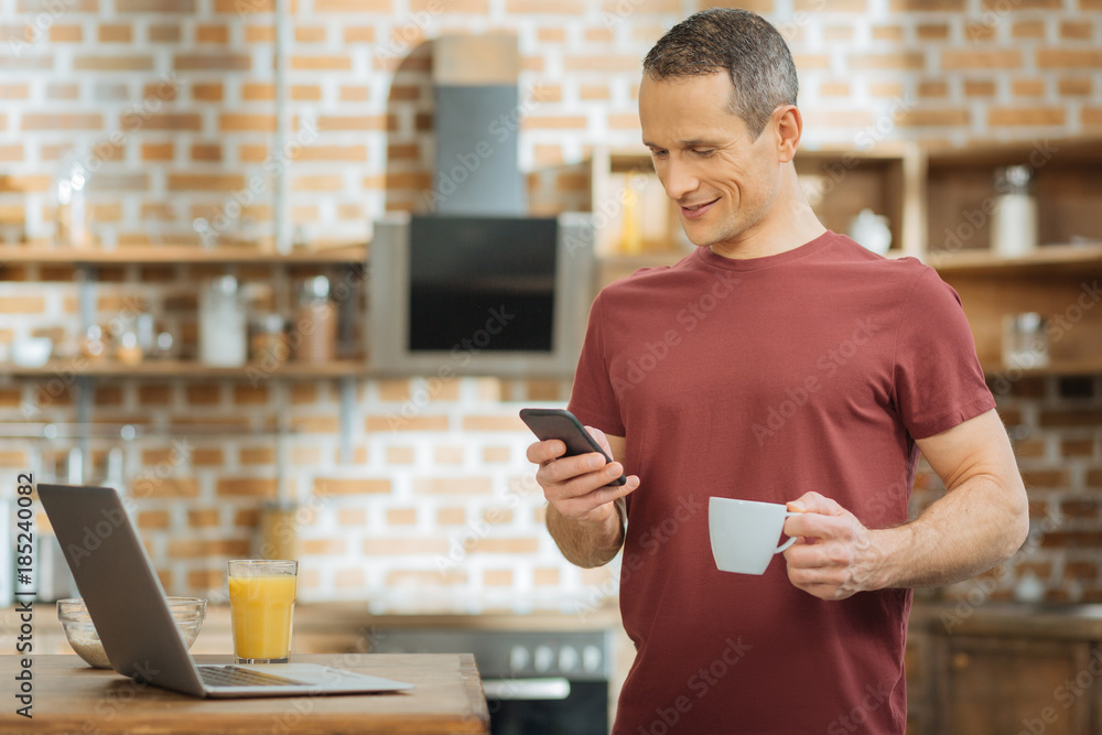 Waiting for you. Attractive male person keeping smile on his face and looking at telephone while standing in the kitchen Waiting for you. Attractive male person keeping smile on his face and looking at telephone while standing in the kitchen