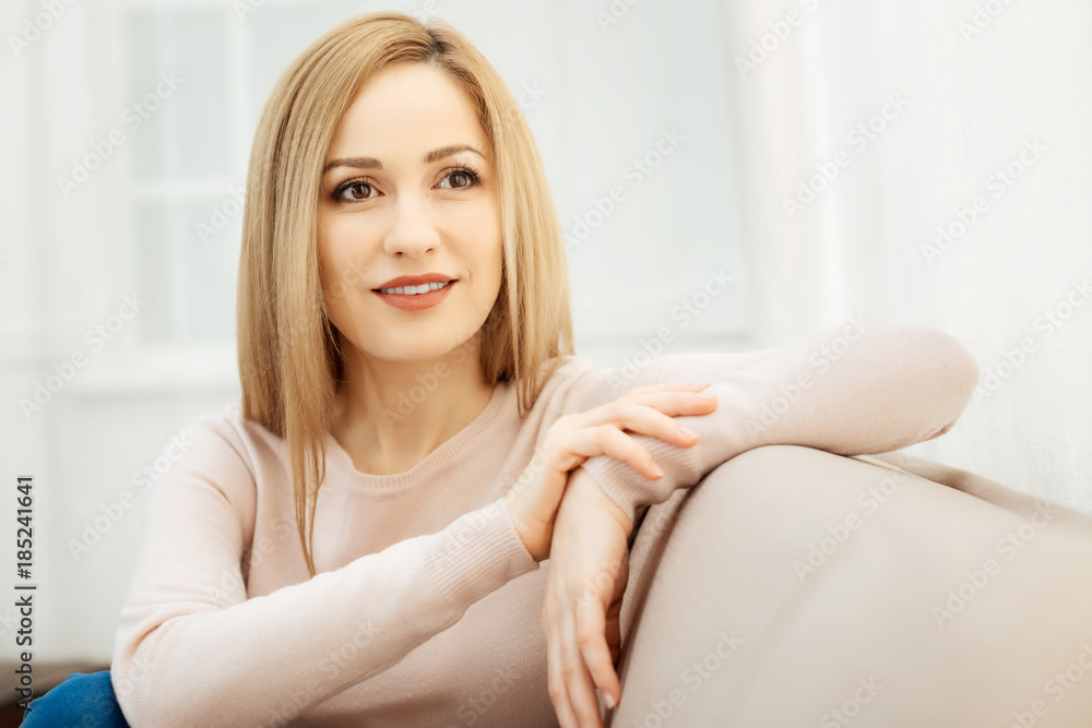 Inspired. Beautiful content young dark-eyed blond woman smiling and wearing a beige sweater and sitting on the couch Inspired. Beautiful content young dark-eyed blond woman smiling and wearing a beige sweater and sitting on the couch
