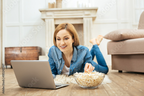 Having fun. Pretty blond smiling long-haired woman lying on the carpet and eating popcorn while using her laptop photo