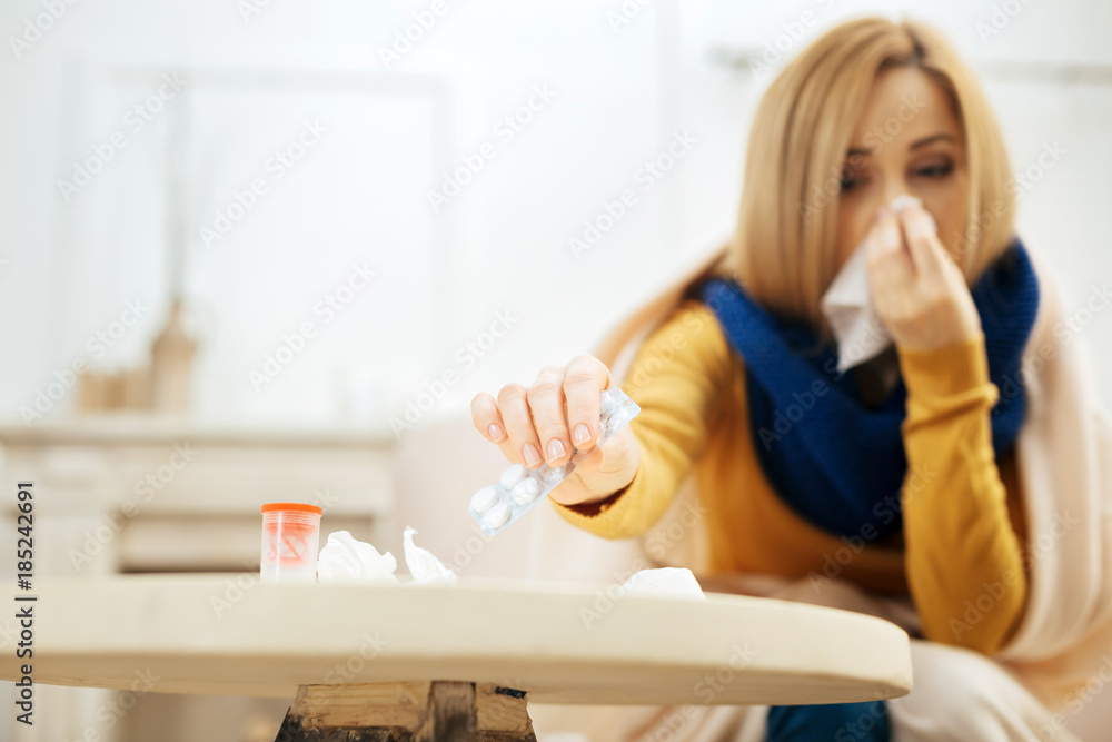 Medications. Sick young blond woman blowing her nose while having a blanket on her shoulders and sitting on the couch and taking the pills from the table in front of her