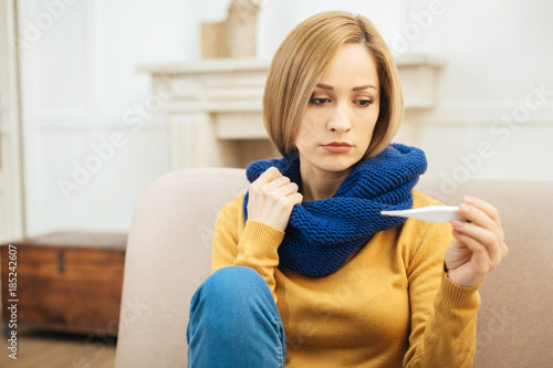 Feeling bad. Beautiful young blond woman feeling down in the mouth and holding a thermometer and having a blue warm scarf on while sitting on the sofa photo