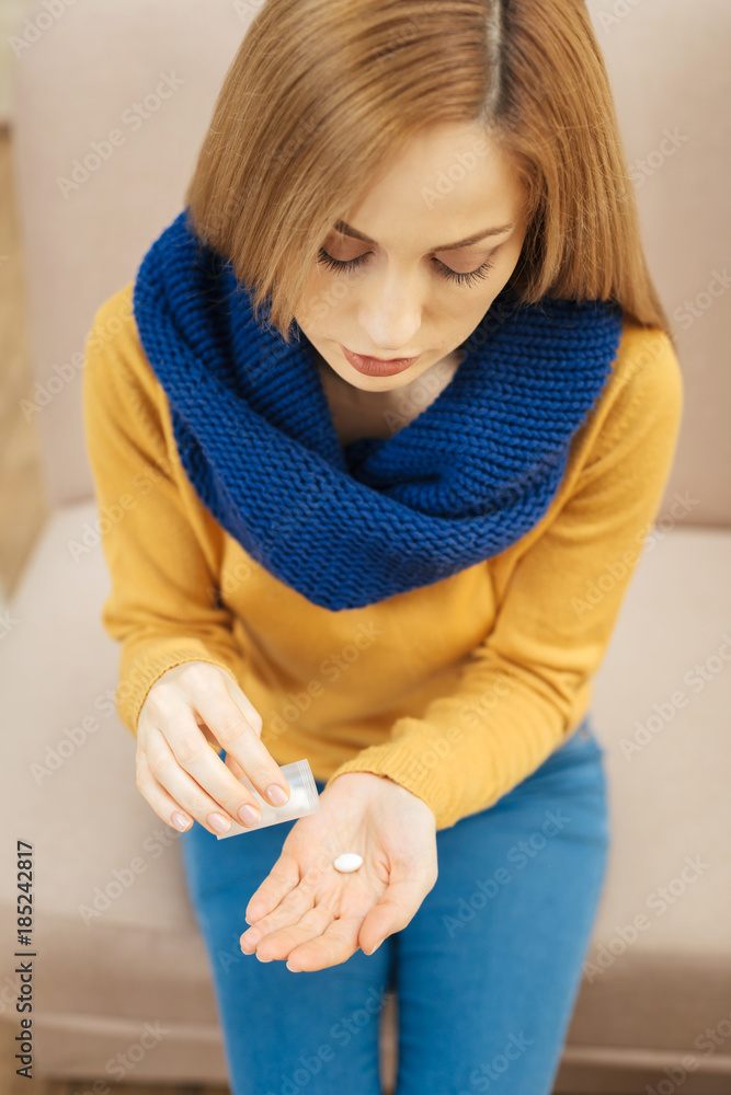 Naklejka premium Recovery. Unhappy young blond long-haired woman wearing a scarf jeans and sweater and sitting on the couch and holding pills
