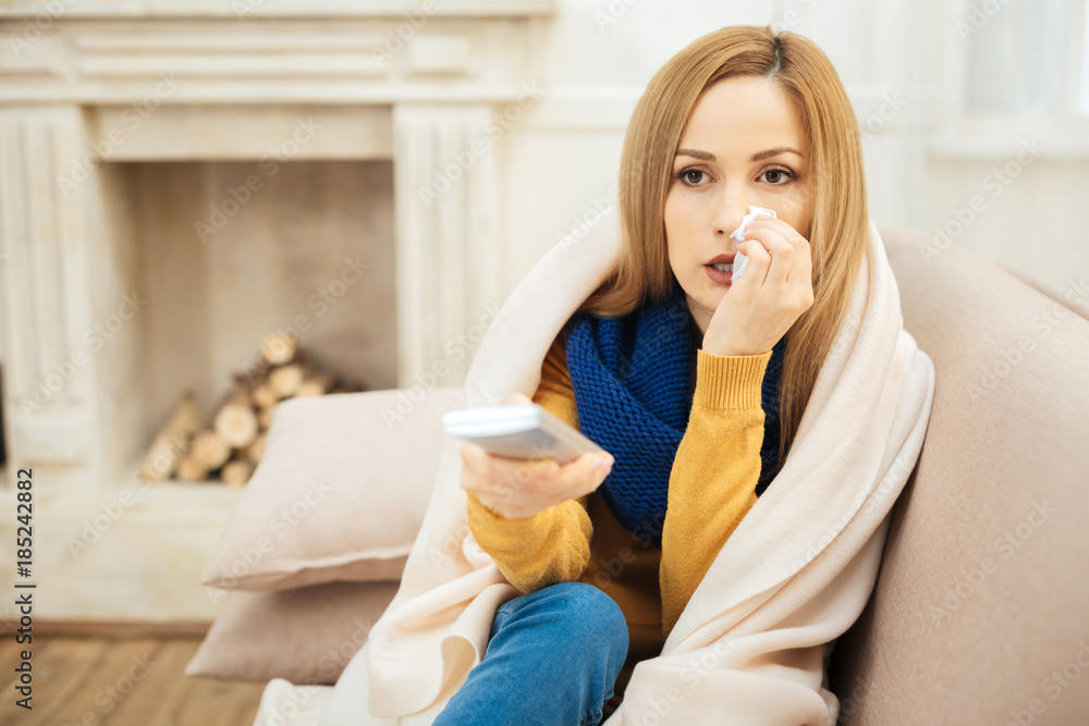 Sick leave. Beautiful cheerless young dark-eyed woman having runny nose and feeling unwell and sitting on the couch and holding remote control Sick leave. Beautiful cheerless young dark-eyed woman having runny nose and feeling unwell and sitting on the couch and holding remote control
