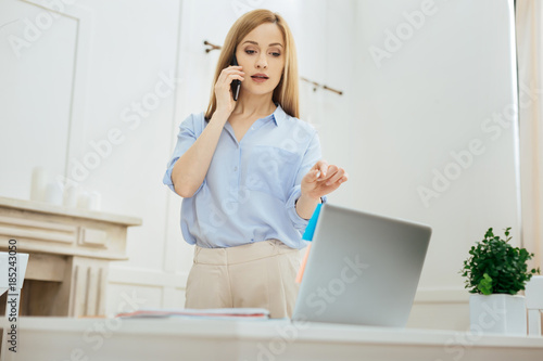 Solving problems. Attractive confident young blond woman wearing a blue shirt and talking over the phone while standing at the table with her laptop and a chimney-piece in the background © Viacheslav Yakobchuk