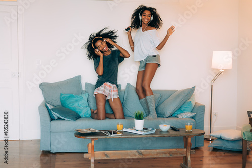 Happy young two black women sitting and standing up in the couch on the computer and mobile phone with headphones dancing .
