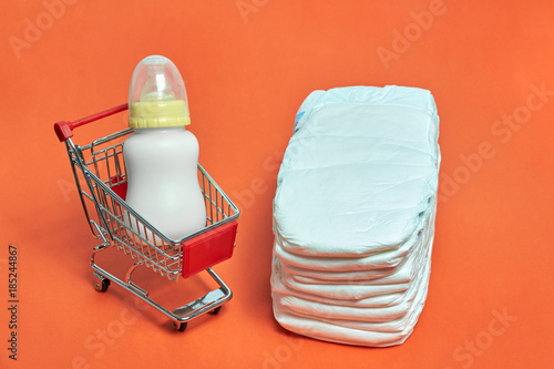 close-up of shopping trolley with baby bottle milk and diappers on orange background with some copy space