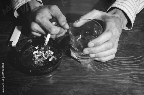Hands of alcoholic man holding a glass with alcohol drink with smoking cigarette in the ashtray black and white