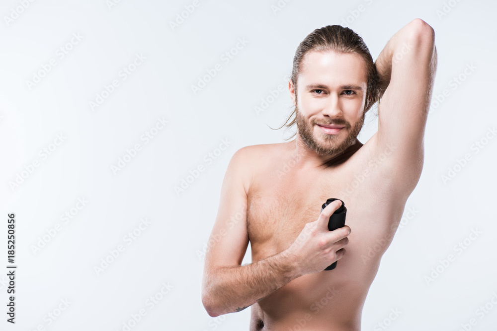 Smiling young man using deodorant spray with hand behind head, isolated on gray Smiling young man using deodorant spray with hand behind head, isolated on gray