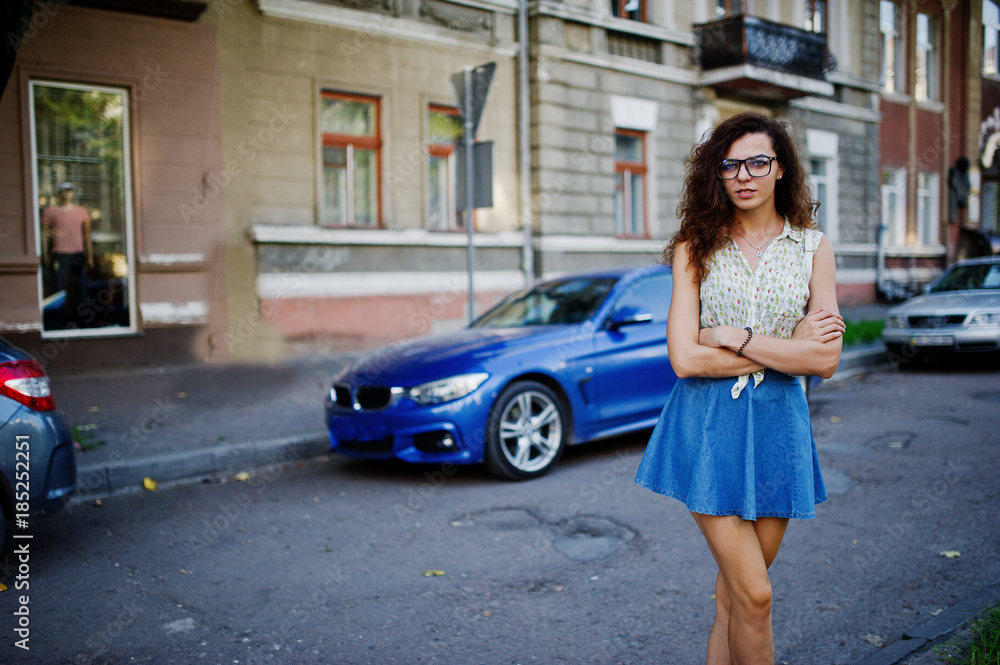 Curly stylish girl wear on blue jeans skirt, blouse and glasses posed near blue car at street of city. Curly stylish girl wear on blue jeans skirt, blouse and glasses posed near blue car at street of city.