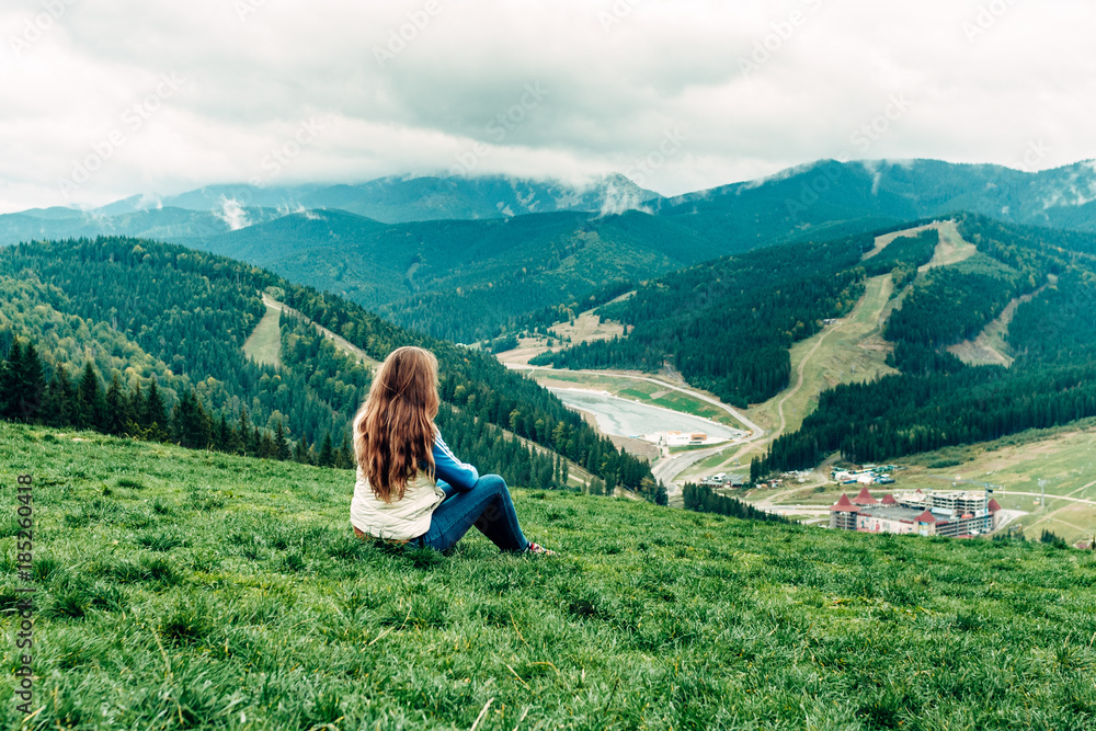 girl, woman sitting sits on a hill and looks down at the beautiful view of the mountains and forests opening in front of her