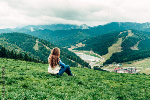 girl, woman sitting sits on a hill and looks down at the beautiful view of the mountains and forests opening in front of her