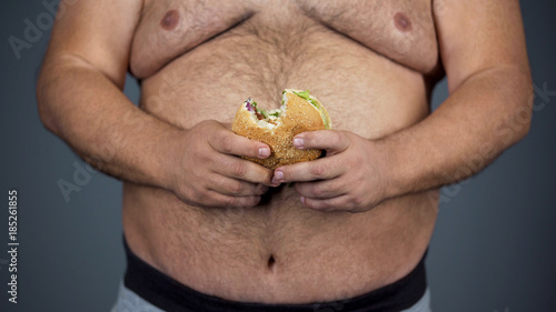 Obese male holding unhealthy hamburger in hands, overweight problem, cholesterol