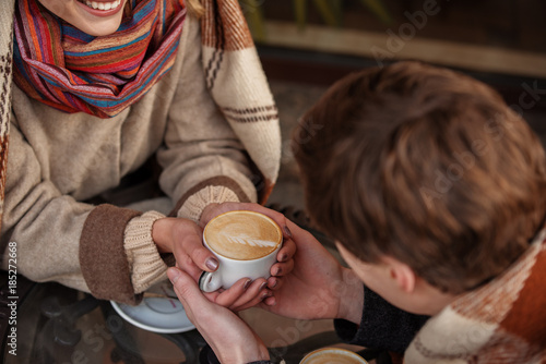 Close up top view of man holding hands of woman while she is having mug of coffee. They are in blankets while sitting at table outdoors