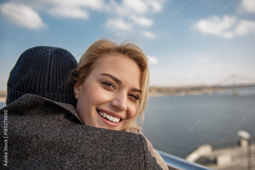 Fototapeta premium Sweet hug. Close up portrait of young woman cuddling man while laughing and looking at camera in open air. Copy space in right side