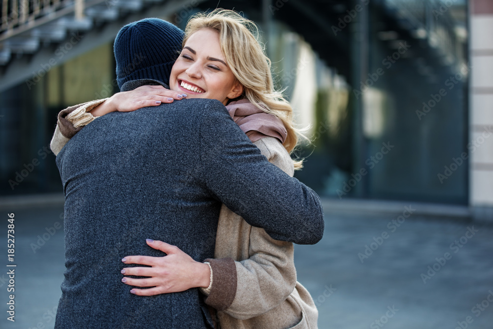 Portrait of smiling woman tightly cuddling man while standing in the street. Lady is with closed eyes while laughing. Copy space Portrait of smiling woman tightly cuddling man while standing in the street. Lady is with closed eyes while laughing. Copy space