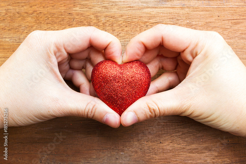Woman hands make a heart shape and holding a glitter red heart decorate object on wooden background.