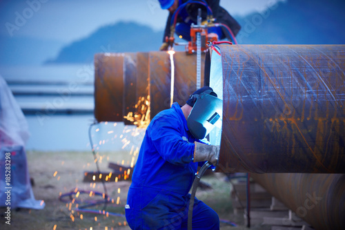 Industrial electrode welder with face shield and blue overall welding a steel pipe in workshop.