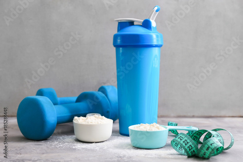 Protein shake in bottle, powder, measuring tape and dumbbells on table