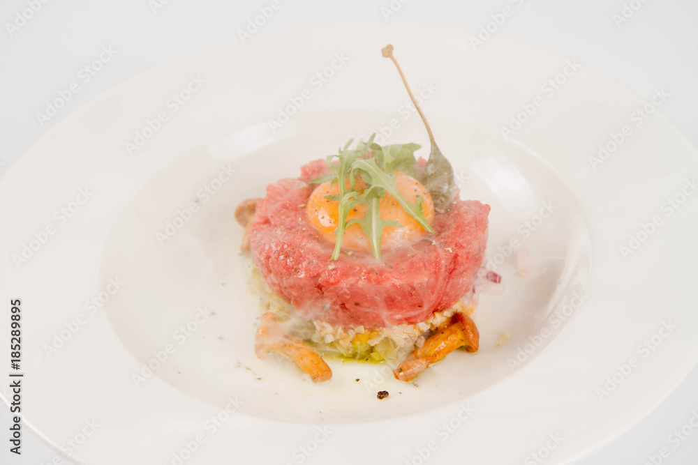 Smoked steak tartare served with egg yolk, onion, pickle, caper, mushroom and rocket salad on a white background Smoked steak tartare served with egg yolk, onion, pickle, caper, mushroom and rocket salad on a white background