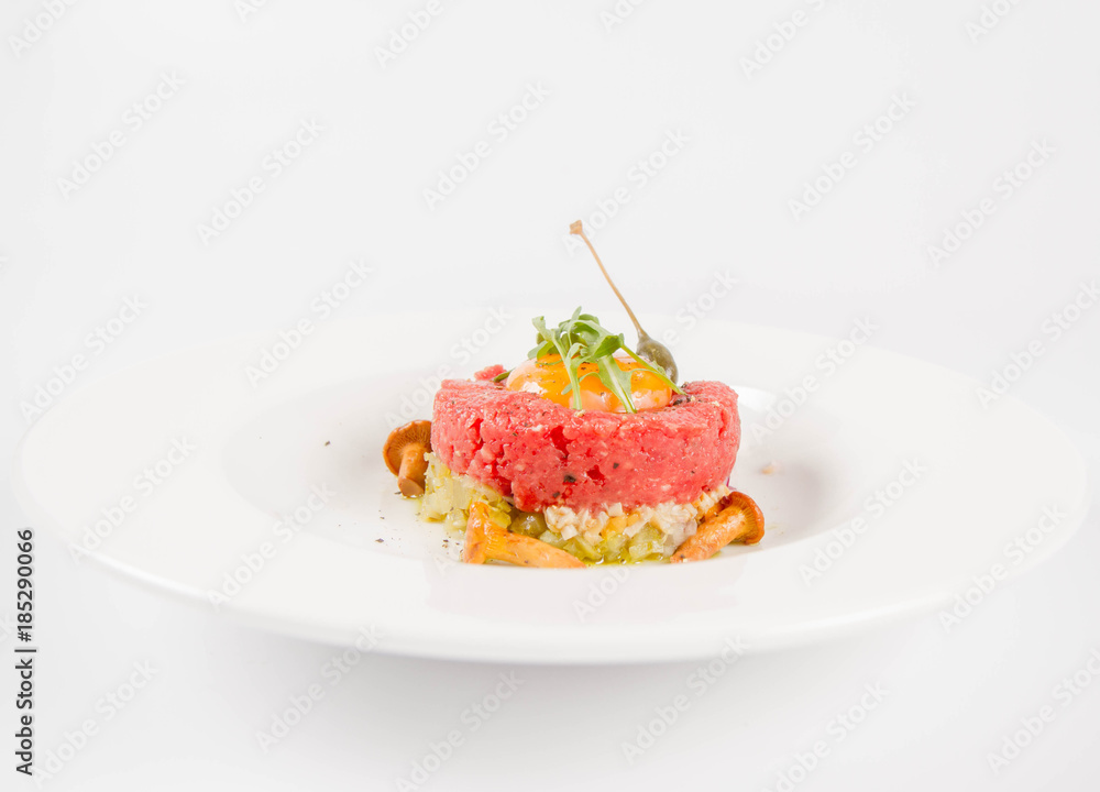 Steak tartare served with egg yolk, onion, pickle, caper, mushroom and rocket salad on a white background Steak tartare served with egg yolk, onion, pickle, caper, mushroom and rocket salad on a white background