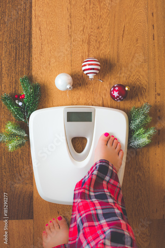 Top view of female feet in winter pajamas on digital scales or weight scale on wooden background surrounded with Christmas lights and decoration. Weight gain during holidays concept, vintage toned