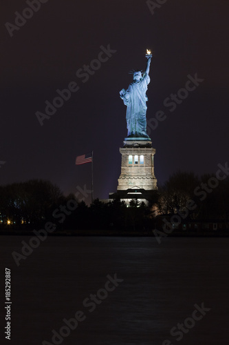 The Statue of Liberty on Ellis Island at night lit by spotlights with an American flag in the wind