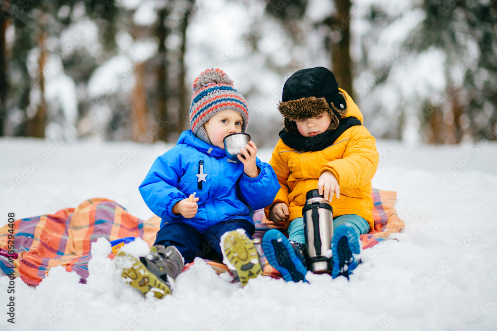 Funny children winter party in snowy forest. Kids male friends rest outdoor at nature. Young boys sharing and drinking tea from thermos. Hot drinks and beverage in cold weather. Warming in frost day.