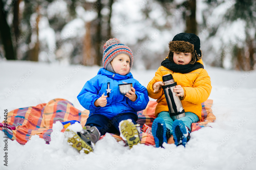 Funny children winter party in snowy forest. Kids male friends rest outdoor at nature. Young boys sharing and drinking tea from thermos. Hot drinks and beverage in cold weather. Warming in frost day.
