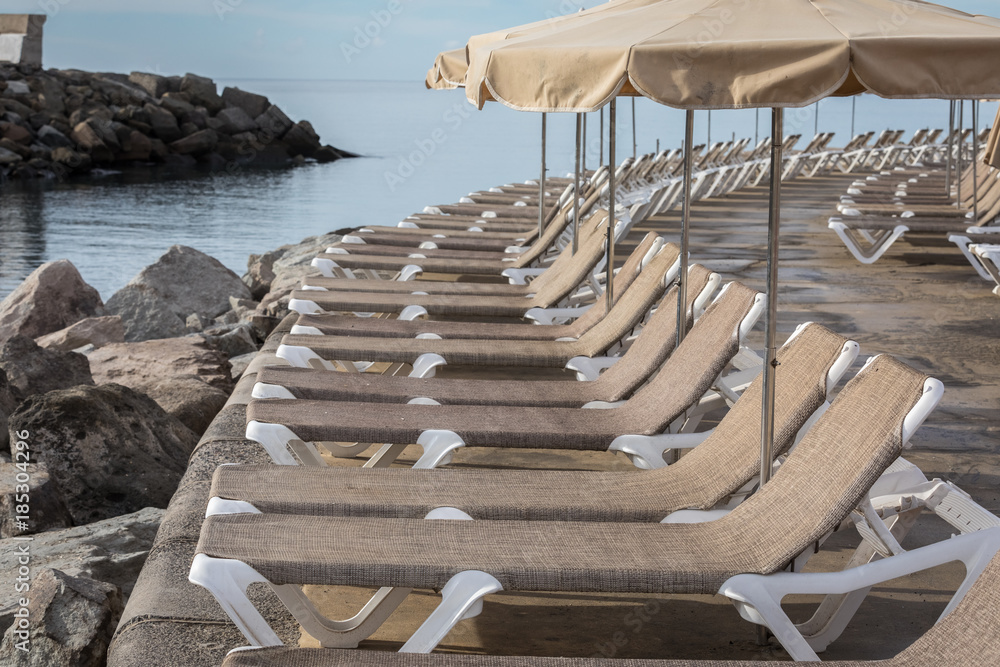 Row of sunbeds and parasols at the breakwater by the beach Playa de Puerto Rico on the Canary Island Row of sunbeds and parasols at the breakwater by the beach Playa de Puerto Rico on the Canary Island