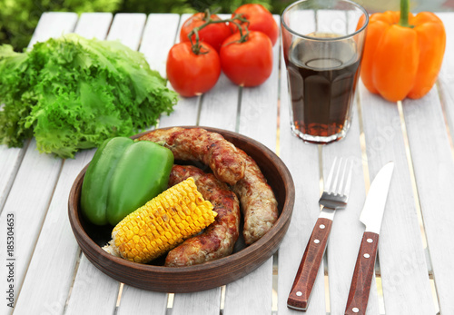 Plate with delicious grilled sausages and vegetables on wooden table outdoors