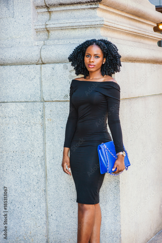 African American Woman street fashion in New York. Wearing long sleeve, slim, off shoulder dress, wristwatch, carrying blue bag, young black girl with braid hairstyle standing by wall, looking at you