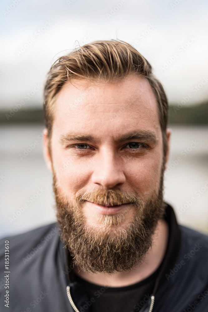 Obraz premium Laid back man with beard standing against a big lake looking at camera outdoors on a sunny day.