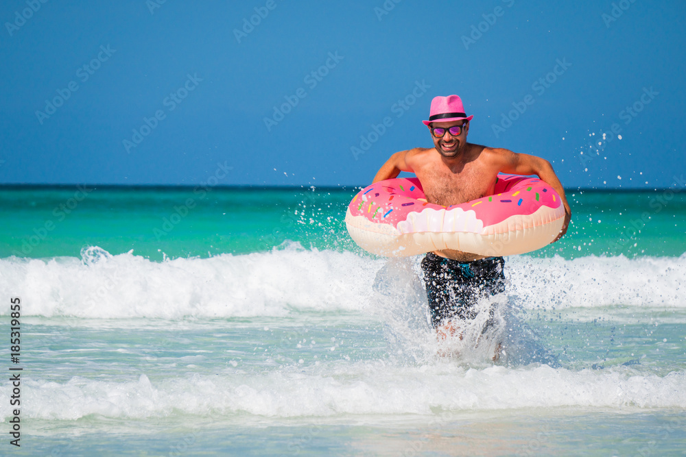 Happy handsome smiling man in pink hat is running with a big inflatable ring on the coast of Caribbean Sea in summer sunny day