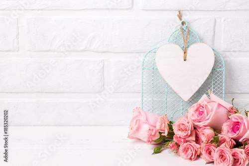 Pink roses flowers and, decorative bird cage and white  heart against  white brick wall.