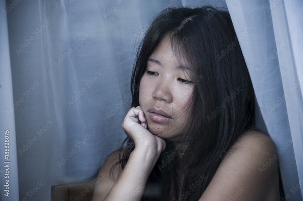 Fototapeta premium young sad and depressed Asian Chinese woman looking thoughtful through window glass suffering pain and depression in sadness concept