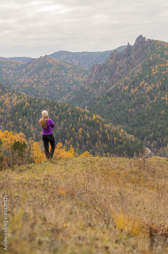 A girl in a lilac jacket looks out into the distance on a mountain, a view of the mountains and an autumnal forest by an overcast day. Free space for text A girl in a lilac jacket looks out into the distance on a mountain, a view of the mountains and an autumnal forest by an overcast day. Free space for text