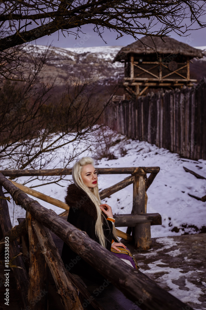 Outdoors portrait of beautiful furious scandinavian warrior ginger woman in a traditional clothes with fur collar, with sword in her hand and wooden Viking Village view on the background. Outdoors portrait of beautiful furious scandinavian warrior ginger woman in a traditional clothes with fur collar, with sword in her hand and wooden Viking Village view on the background.