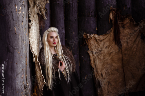 Outdoors portrait of beautiful furious scandinavian warrior ginger woman in a traditional clothes with fur collar  with sword in her hand and wooden Viking Village view on the background.