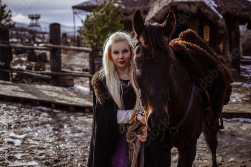 Outdoors portrait of beautiful furious scandinavian warrior ginger woman in a traditional clothes with fur collar, with sword in her hand and wooden Viking Village view on the background.