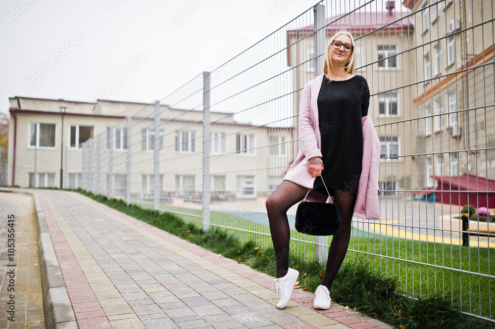 Blonde girl at glasses and pink coat, black tunic and handbag posed against fence at street. Blonde girl at glasses and pink coat, black tunic and handbag posed against fence at street.