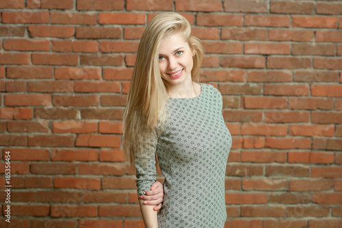 Portrait of a blonde in a light sweater. Standing against the red brick wall background, smiling and posing for the camera. her long hair is laid in one direction © alexsfoto