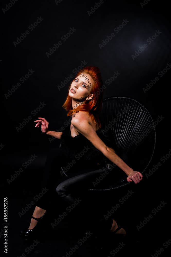 Fashion model red haired girl with originally make up like leopard predator isolated on black. Studio portrait on chair. Fashion model red haired girl with originally make up like leopard predator isolated on black. Studio portrait on chair.