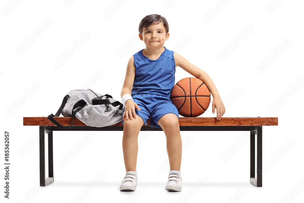 Obraz premium Little basketball player sitting on a wooden bench next to a sports bag