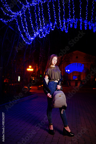 Night portrait of girl model wear on glasses  jeans and leather jacket  with backpack in hands  against blue lights garland of city street.